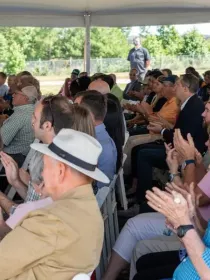 A large group of people seated under a tent, clapping and facing forward. Some individuals wear hats and sunglasses. Trees and a fence are visible in the background.