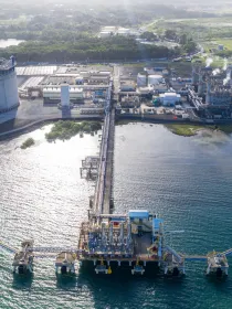 Aerial view of a large industrial complex by the water, featuring a cylindrical storage tank, processing facilities, and docks extending into the sea.