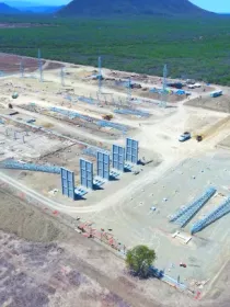 Aerial view of a large construction site in a rural area with several steel structures and towers being assembled. The site is surrounded by open land and distant hills under a clear blue sky.