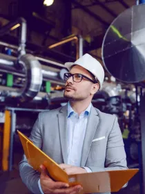 A man in a gray suit and white hard hat holds a folder while inspecting an industrial facility with large metal pipes and machinery.