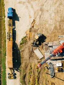 Aerial view of a construction site with a blue truck connected to a long yellow trailer on a dirt road. Nearby, a red crane is positioned on wooden planks, and a bulldozer is visible on the dirt. Adjacent to the site is a green field.