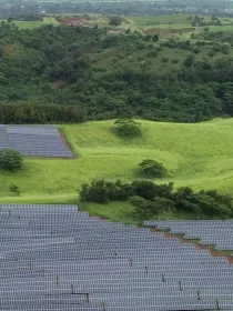 Aerial view of a solar farm with numerous solar panels arranged in rows on a lush green hillside. The landscape is surrounded by dense trees and vegetation, with a distant view of more hills and a town in the background.