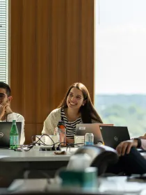 A group of people sitting around a conference table with laptops and bottles. A woman in the center smiles, while others listen attentively. One person wears a face mask.