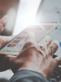 Close-up of hands holding a smartphone with a laptop and financial graphs in the background.