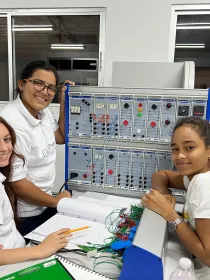 Three women in a classroom setting work together on an electrical engineering project. They are smiling and wearing white shirts with logos. A panel with various controls and wires is in front of them, and one woman is holding a notebook and pencil.