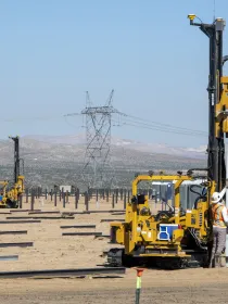 Construction workers operate heavy machinery in a desert landscape, installing metal poles into the ground. The scene includes several workers in safety vests and helmets, with mountains and power lines visible in the background under a clear blue sky.
