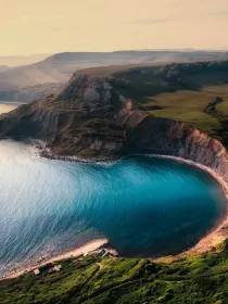 Aerial view of a coastal landscape with steep cliffs and a curved bay with turquoise water. The cliffs are covered in green vegetation, and the sea reflects the light of the setting sun.