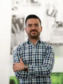 A person with short dark hair and glasses stands with arms crossed, wearing a blue and white checkered shirt. The background is blurred, featuring abstract black and white patterns.