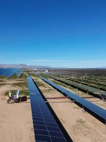 A vast solar panel farm under a clear blue sky, with rows of solar panels stretching into the distance. Two workers in yellow vests stand near equipment on a dirt path in the foreground. Mountains are visible on the horizon.