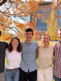 Five people standing closely together, smiling under a tree with autumn leaves. A modern brick building and mountains are visible in the background.