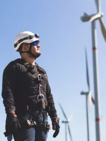 A worker wearing safety gear and a helmet stands in front of wind turbines on a clear day.