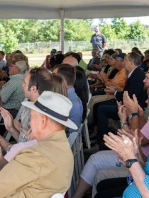 A diverse group of people seated under a tent, attentively watching an event outdoors. Many are clapping, wearing casual and semi-formal attire. The background shows trees and a fence.