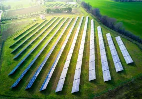 Aerial view of a solar farm with multiple rows of solar panels on a grassy field surrounded by trees and a road.