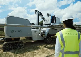 A person wearing a safety vest and hard hat observes a solar panel installation robot labeled 'maximo' on a sunny day.