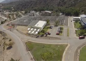 Aerial view of an industrial facility labeled 'AES Gener' surrounded by roads and greenery. The facility includes a main building, parking lot with several cars, and adjacent structures. Nearby, there is a substation with electrical equipment and power lines. The area is set in a mountainous landscape with trees and winding roads.