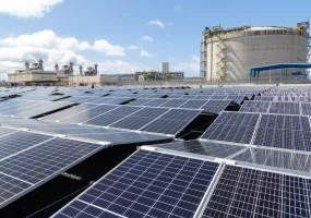 A large field of solar panels under a blue sky, with an industrial facility and a large cylindrical structure in the background.