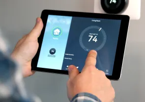 Person adjusting a smart thermostat using a tablet. The screen shows a temperature setting of 74 degrees for the living room. A wall-mounted thermostat displays the same temperature.