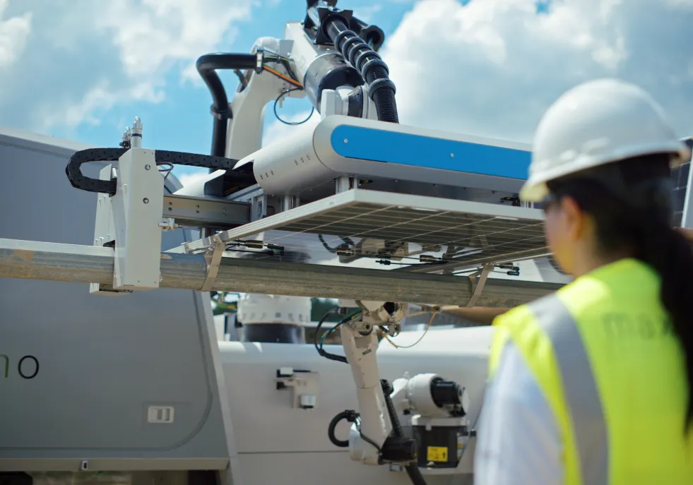A worker in a hard hat and high-visibility vest observes a robotic arm installing a solar panel under a blue sky.