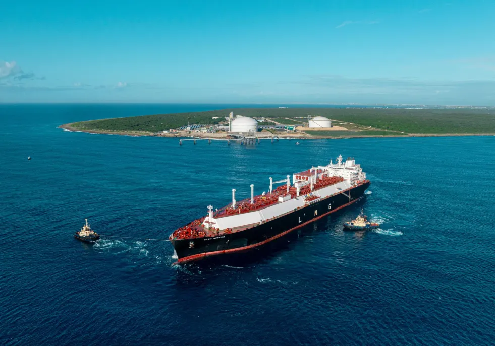 A large LNG tanker is escorted by two tugboats on a vast blue ocean. In the background, an island with green vegetation and white storage tanks is visible under a clear blue sky.