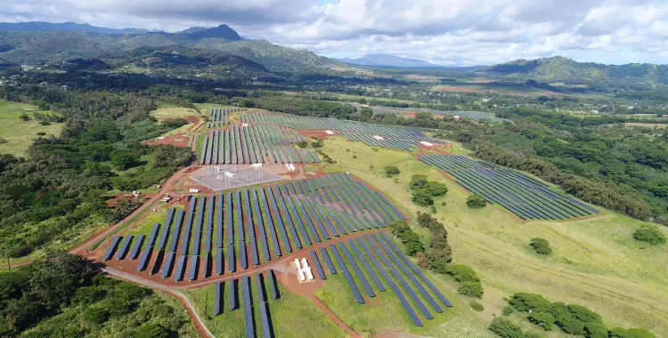 Aerial view of a large solar farm with numerous rows of solar panels set on green grassy fields surrounded by trees and hills under a partly cloudy sky.