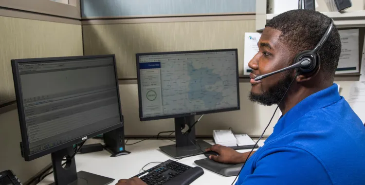 Man in a blue shirt wearing a headset, working at a desk with two computer monitors displaying data and maps in an office cubicle.