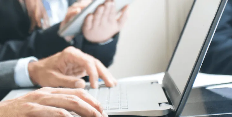 Close-up of hands typing on a laptop keyboard, with another person holding a tablet in the background.
