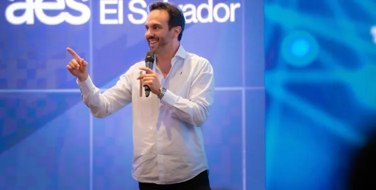 A man in a white shirt speaks into a microphone while gesturing with his hand on stage. The background displays the text 'aes El Salvador' on a large screen.