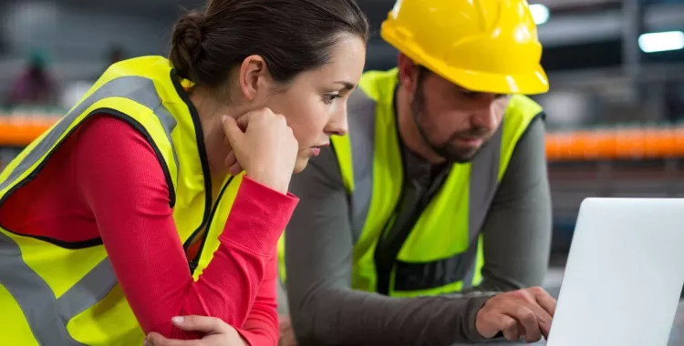 A woman and a man in high-visibility vests are working together at a laptop in an industrial setting. The man is wearing a yellow hard hat. Both are focused on the screen.