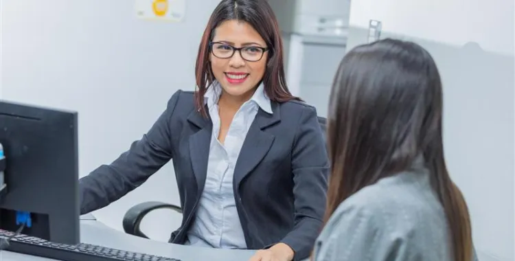 A woman in a business suit and glasses sits at a desk, smiling and talking to another person. A computer and documents are on the desk.