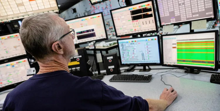 A person in a control room monitors multiple computer screens displaying complex data and graphs. The screens show various charts and colorful diagrams, indicating a high-tech environment.