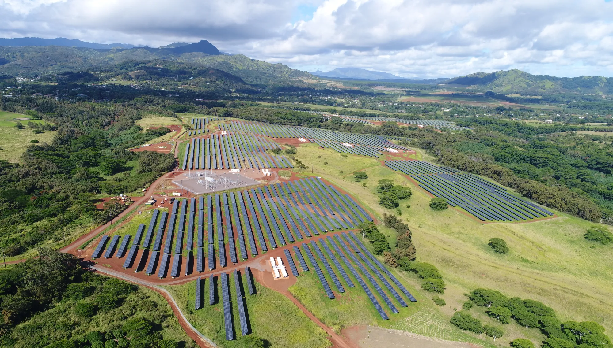 Aerial view of a large solar farm with rows of solar panels on a grassy landscape, surrounded by trees and distant mountains under a partly cloudy sky.