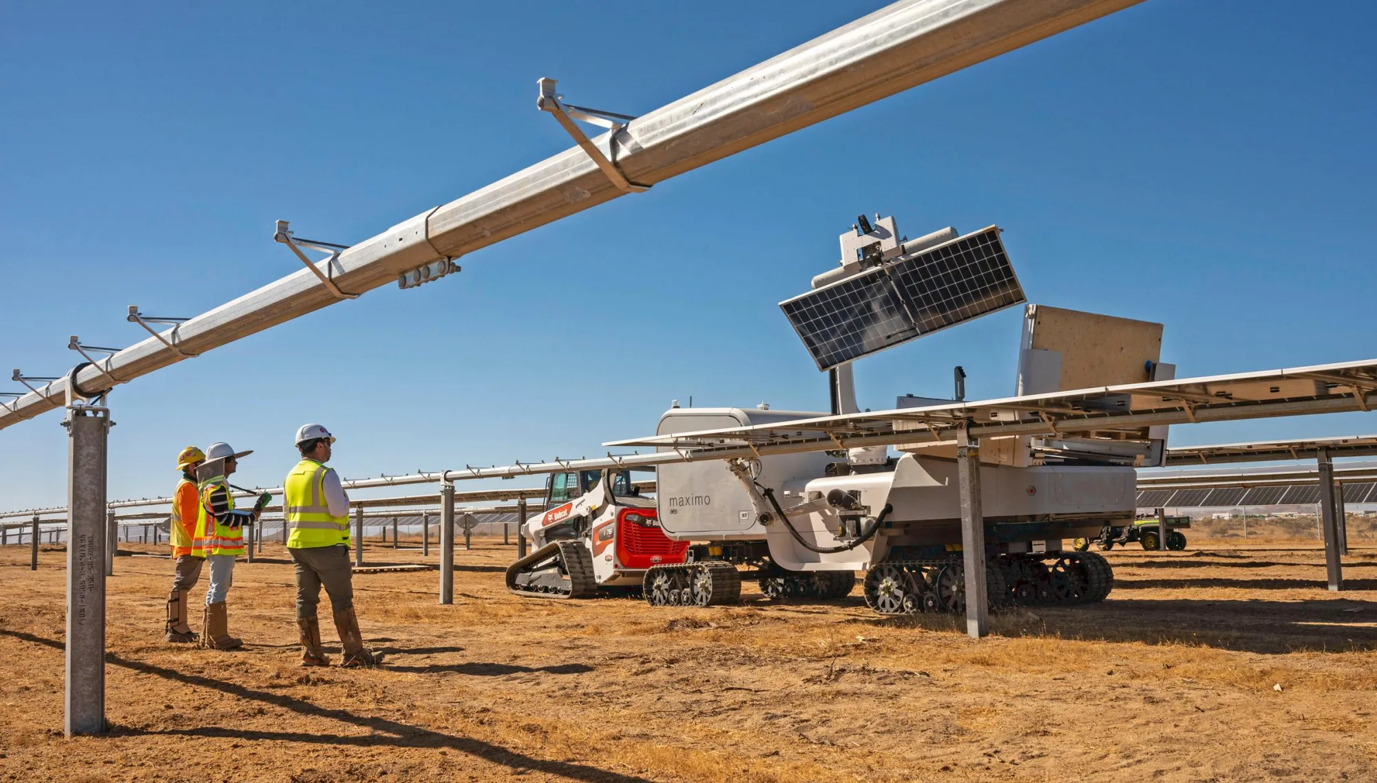 Three workers in safety gear stand beside a solar panel installation in a dry, open field. A robotic machine with tracks is positioned under the panels, likely for maintenance or cleaning. The sky is clear and blue.