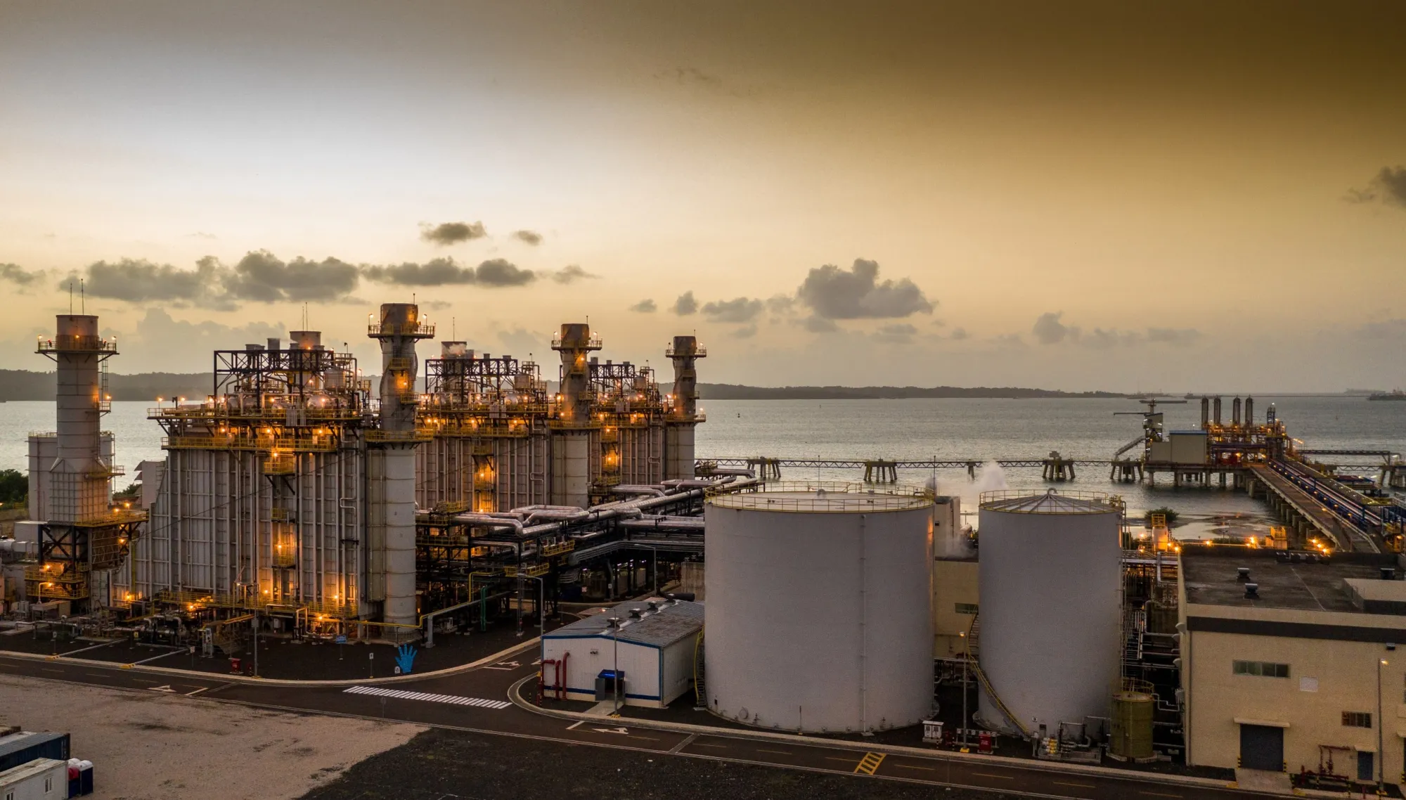 A large industrial power plant with multiple chimneys and storage tanks near a waterfront at sunset. The sky is partly cloudy, and the facility is illuminated with lights.
