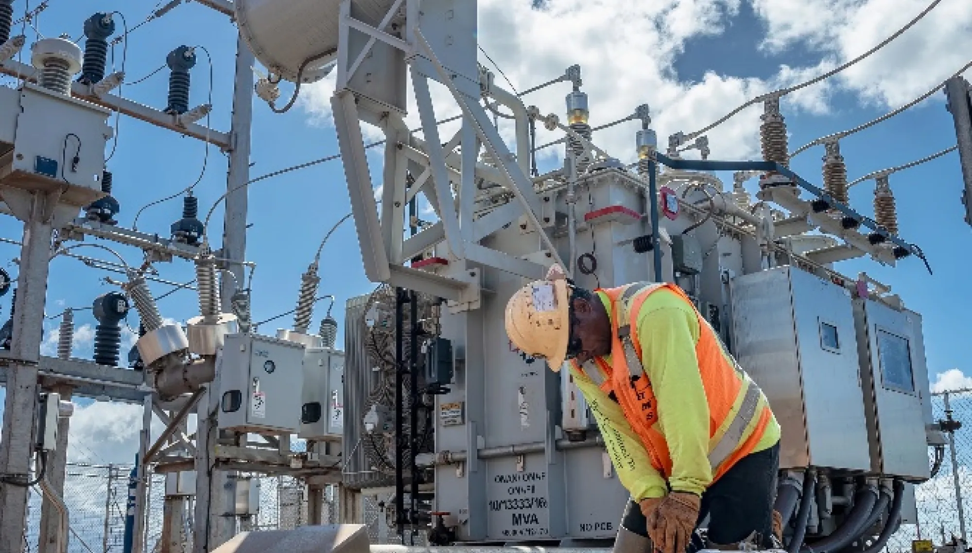 A worker in a safety vest and helmet kneels in front of electrical equipment at a power substation. The sky is partly cloudy in the background.