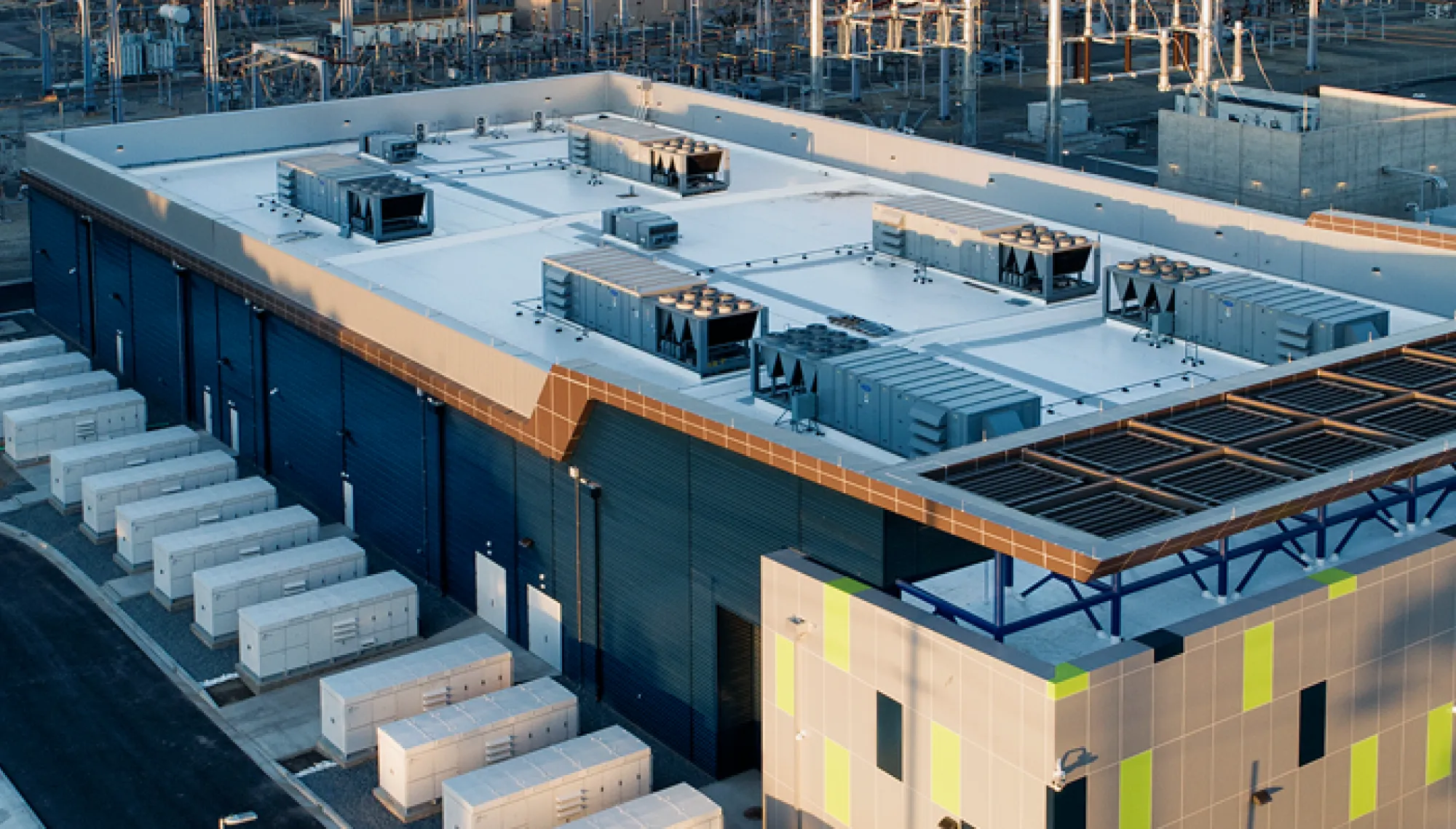 Aerial view of a large industrial facility with a flat roof, featuring multiple HVAC units. The building has a modern design with blue and green accents. Surrounding the facility are several smaller rectangular structures and a network of power lines and equipment, indicating an energy-related site.