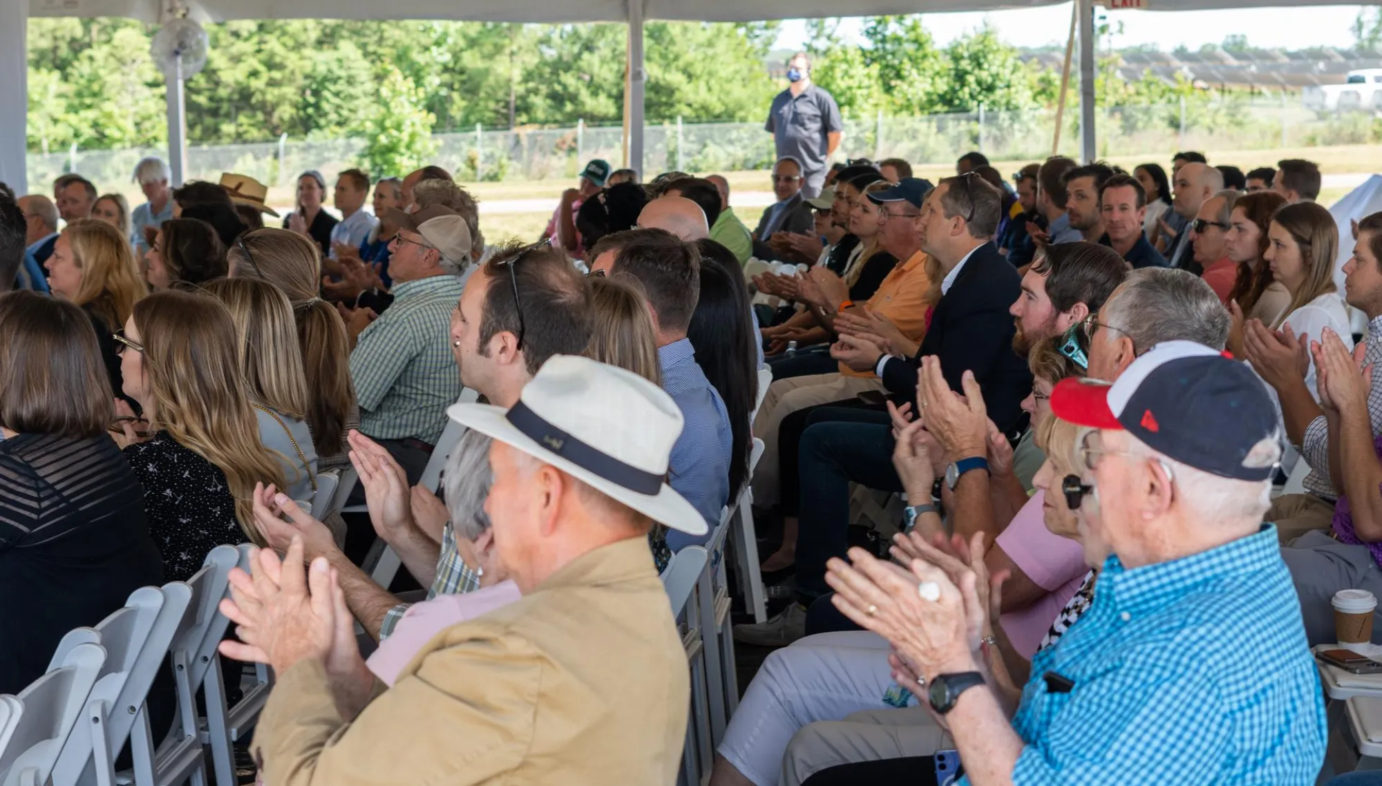 A diverse group of people seated under a tent, attentively watching an event and clapping. The setting appears to be outdoors with trees visible in the background.
