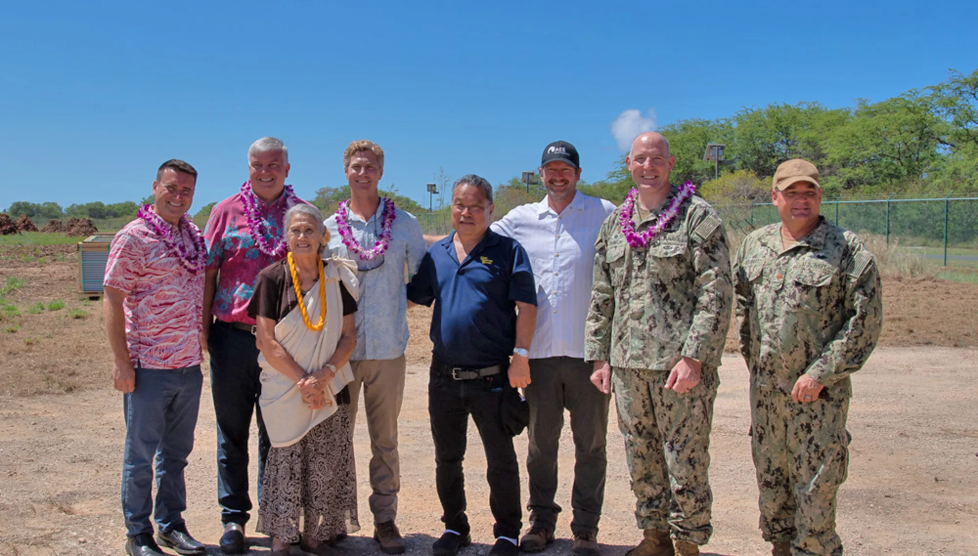 A group of eight people stands outdoors on a sunny day. Some are wearing military uniforms, while others wear casual or Hawaiian-style clothing with leis around their necks. They are standing on a dirt path with trees and a clear blue sky in the background.