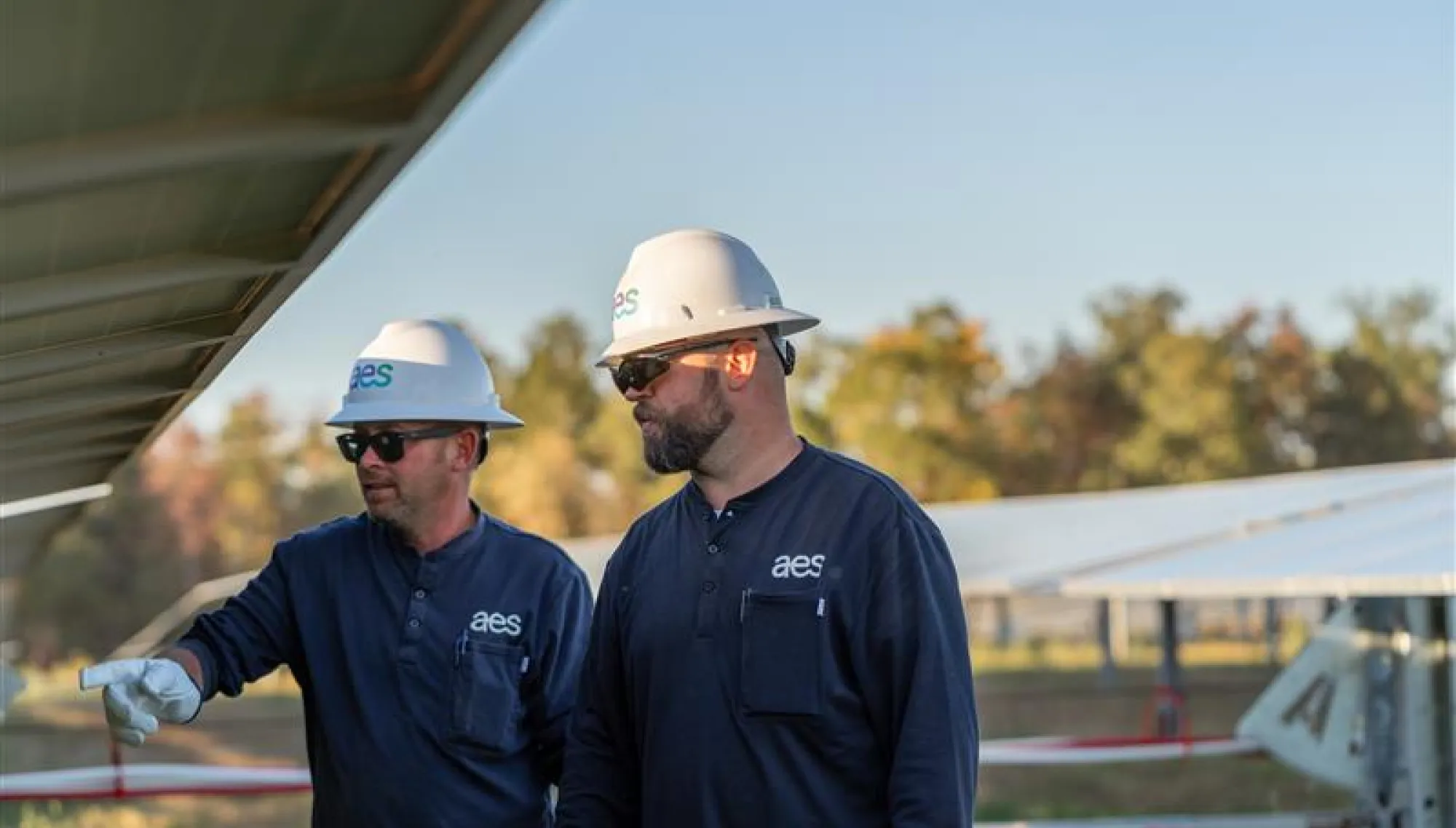 Two workers wearing AES-branded hard hats and shirts inspect solar panels at a solar farm. They are standing outdoors with trees and additional solar panels in the background.