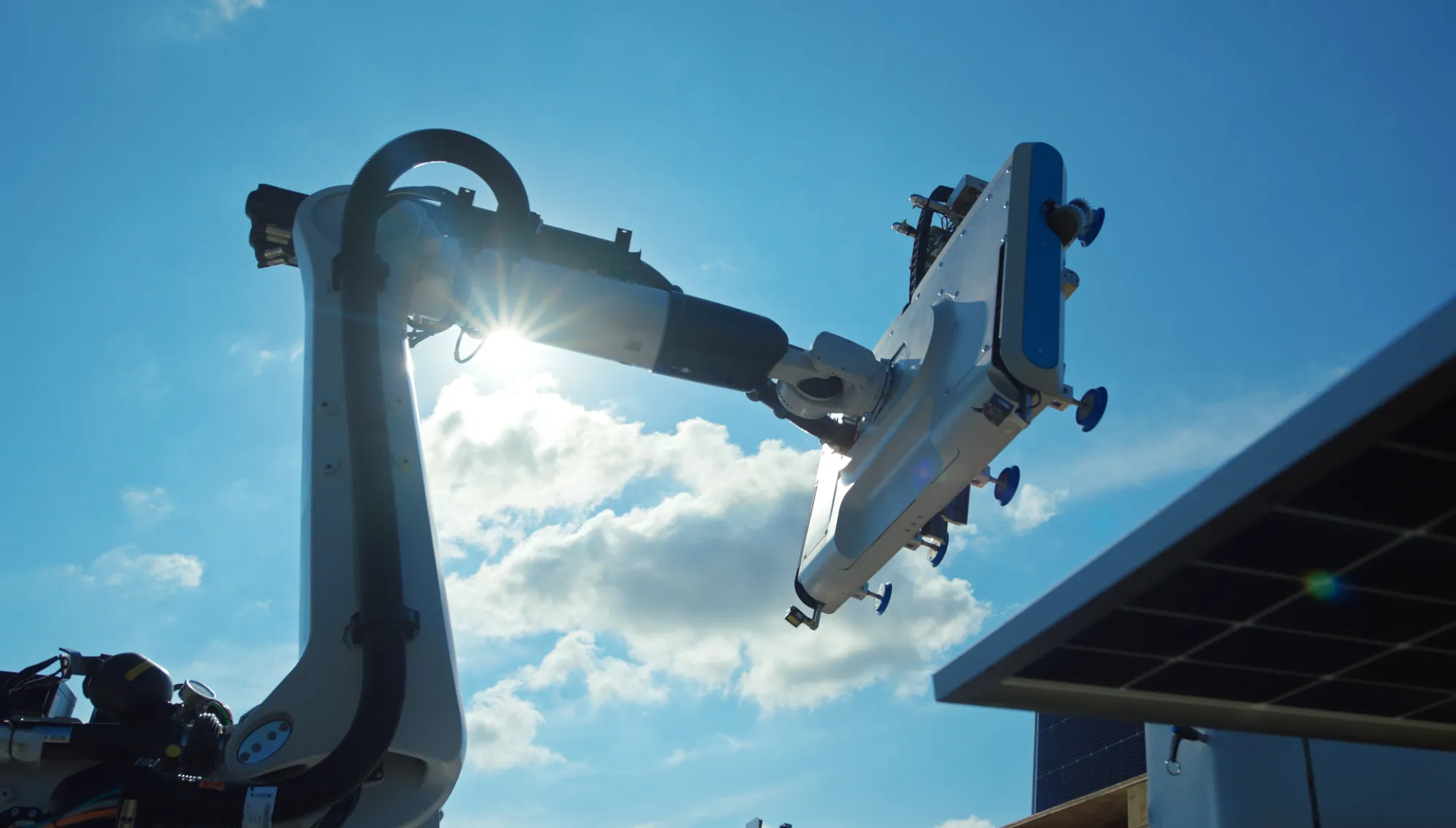 A robotic arm equipped with a large panel is positioned against a bright blue sky with scattered clouds. Sunlight shines through the arm's joints, creating a lens flare. Part of a solar panel is visible in the lower right corner.