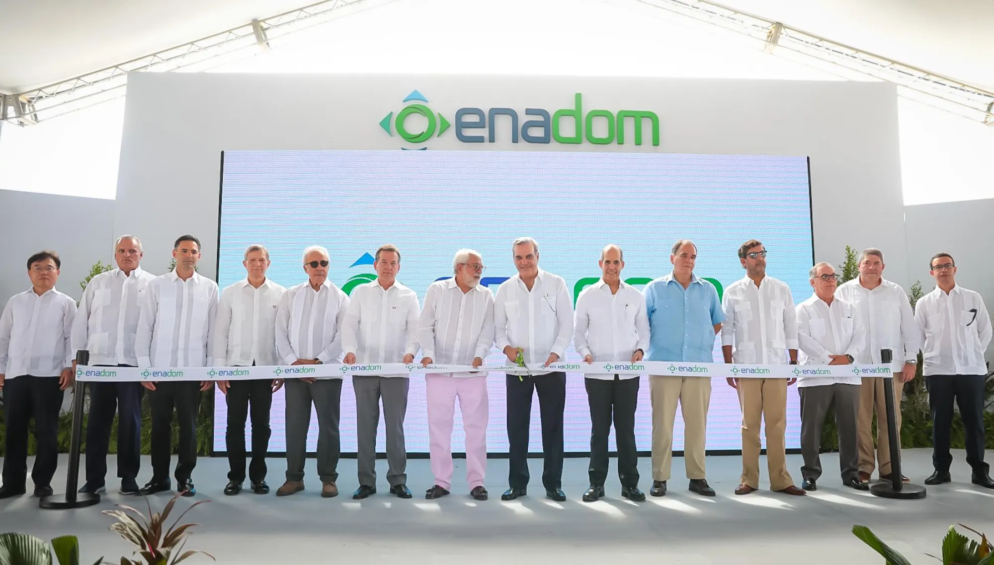 A group of people in white shirts stands in front of a large Enadom sign during a ribbon-cutting ceremony.