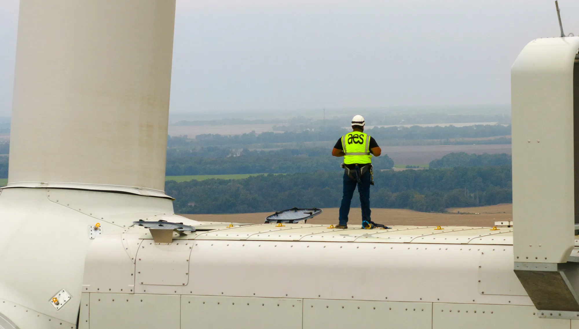 A person wearing a safety harness and a helmet stands on top of a wind turbine. They are wearing a high-visibility vest with 'aes' printed on the back. In the background, there is a landscape of fields and trees under a cloudy sky.