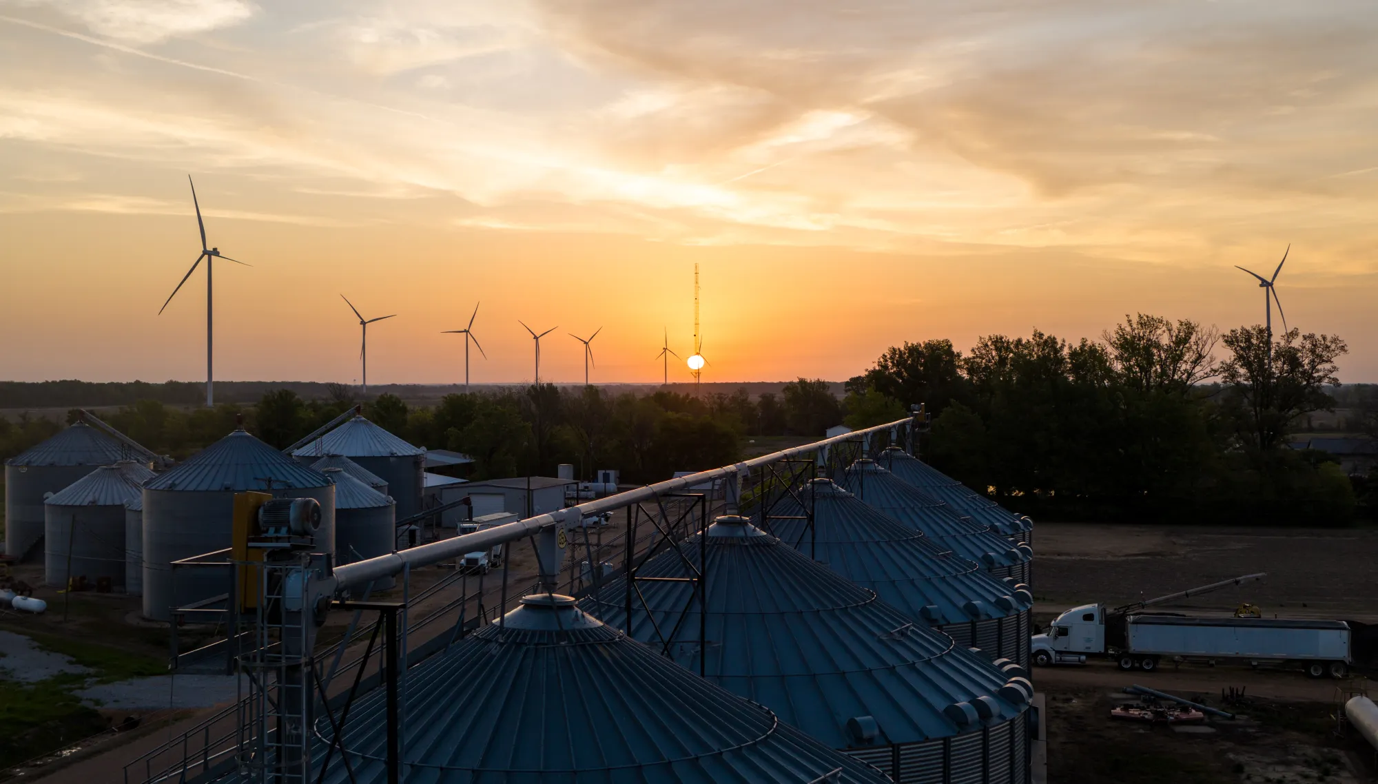 A sunset over a rural landscape with wind turbines in the background. In the foreground, several large metal silos are visible, with a truck parked nearby. Trees and open fields surround the area, and the sky is filled with warm hues of orange and yellow.