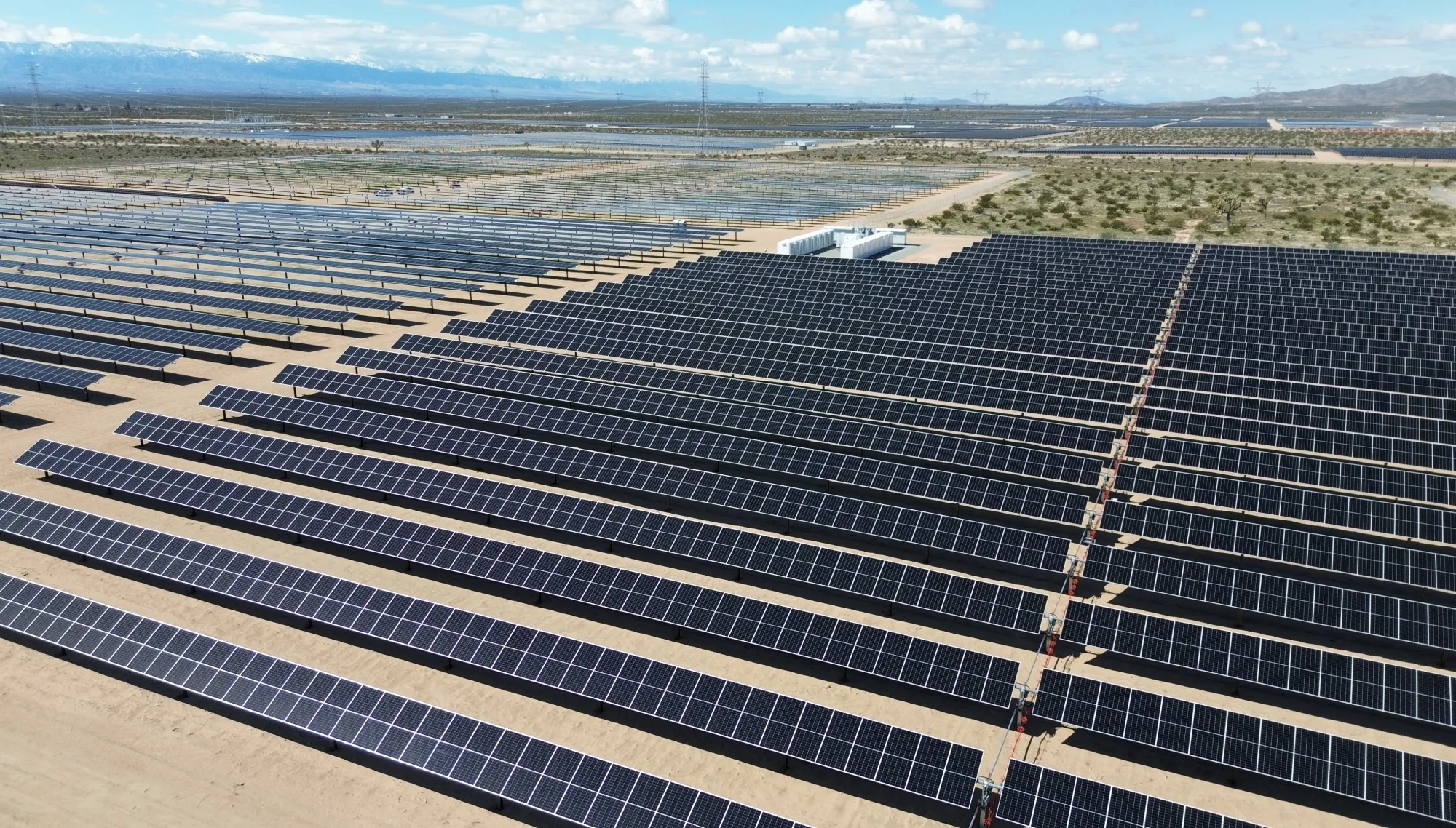 Aerial view of a large solar farm with rows of solar panels stretching across a desert landscape. The panels are aligned in neat rows, and a few small buildings are visible in the distance. The sky is partly cloudy, and mountains can be seen on the horizon.