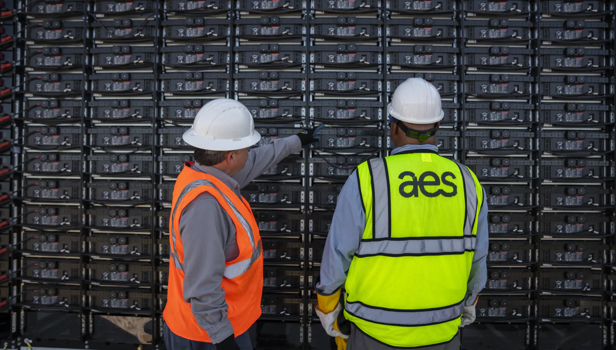 Two workers wearing hard hats and safety vests stand in front of a large wall of battery units. One worker points at the batteries, while the other, wearing a vest with the AES logo, observes.