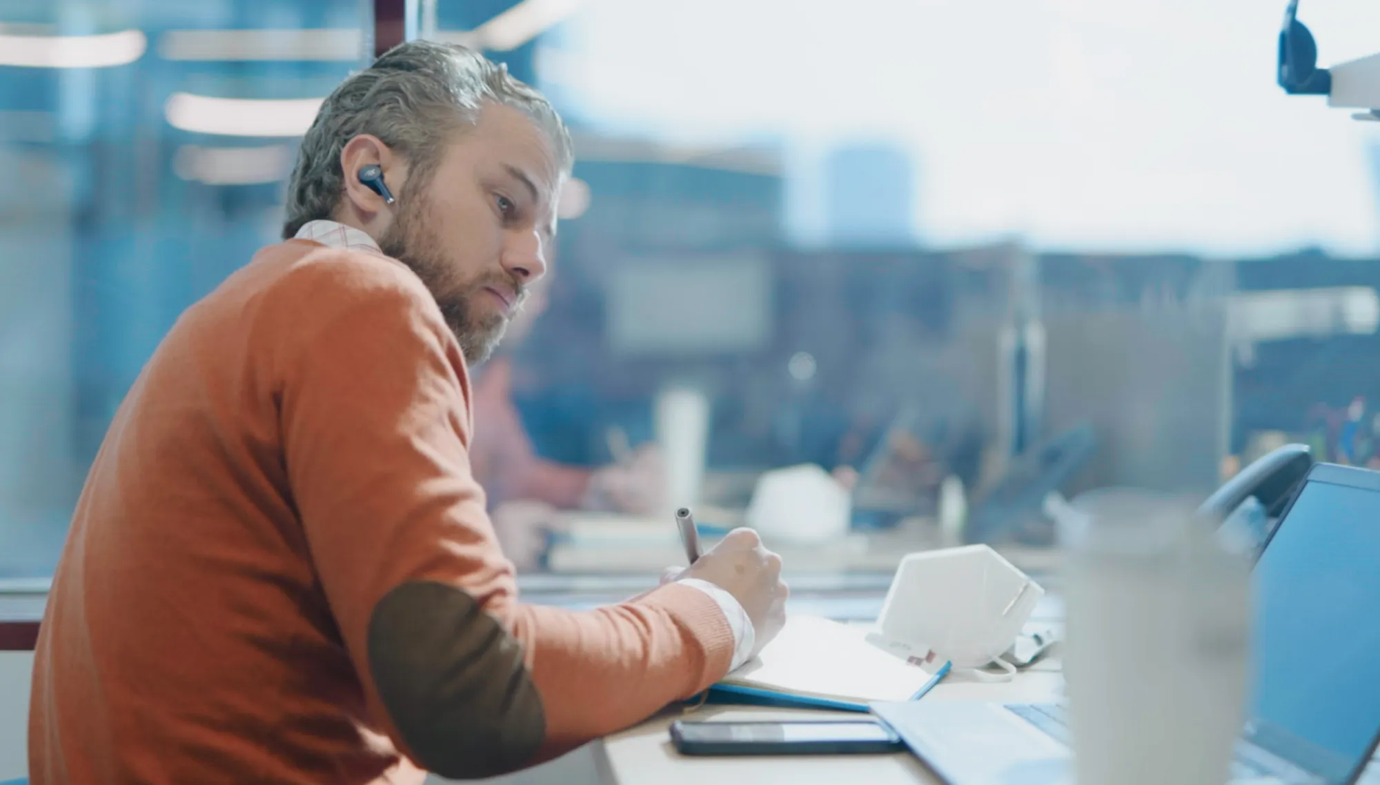 A man with earphones sits at a desk writing in a notebook, with a laptop and a smartphone nearby. The background shows a blurred office setting.