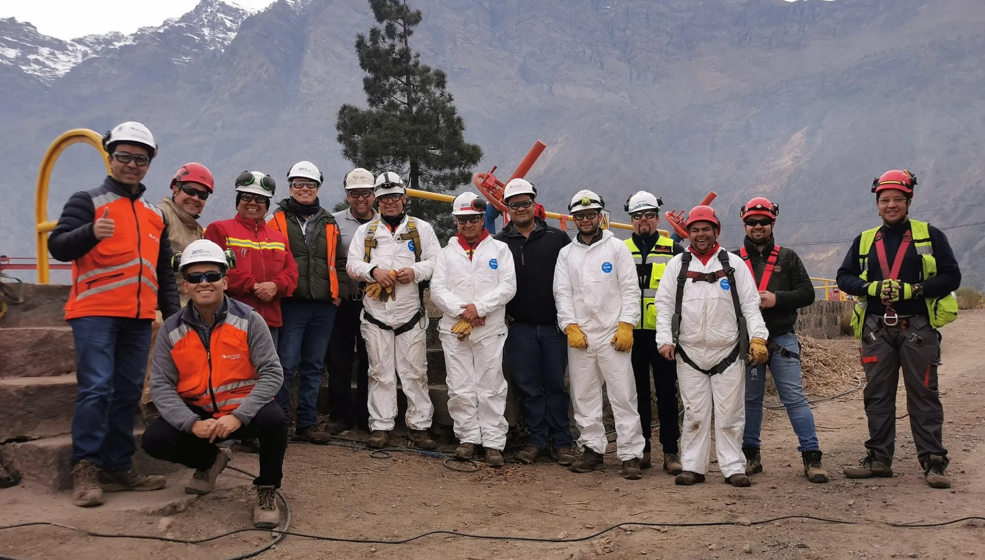 A group of construction workers in safety gear, including helmets and reflective vests, pose for a photo outdoors with a mountainous landscape in the background.