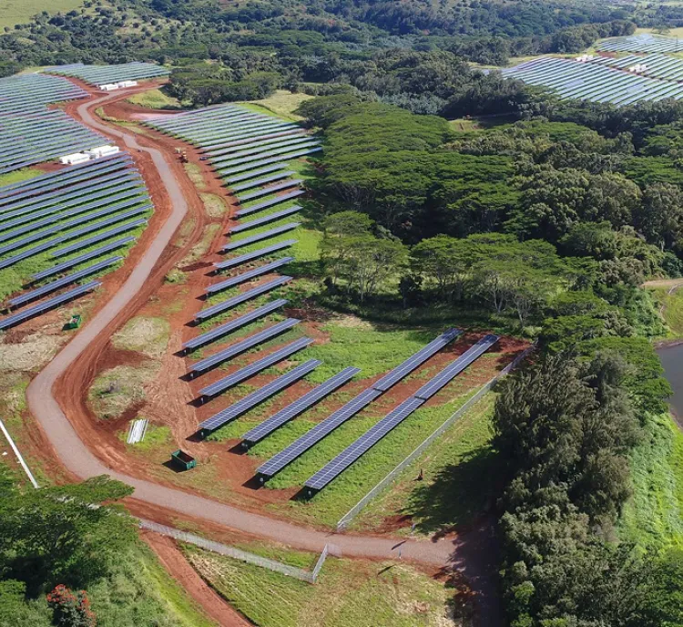Aerial view of a large solar farm with rows of solar panels on red soil, surrounded by lush green trees and vegetation.