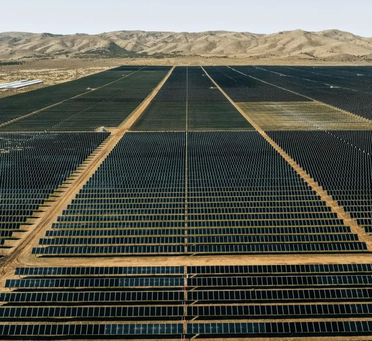 Aerial view of an extensive solar farm with rows of solar panels stretching into the distance, set against a backdrop of arid hills and a clear sky.