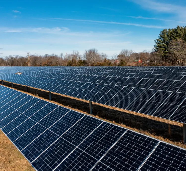 Aerial view of a large solar farm with rows of solar panels under a partly cloudy sky, surrounded by trees.