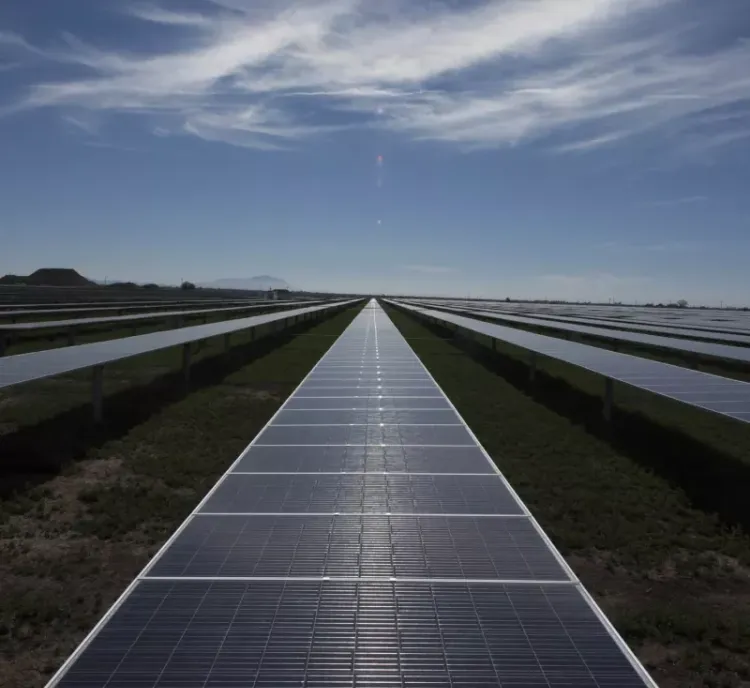 A vast field of solar panels under a clear blue sky with scattered clouds. The panels are aligned in rows and stretch into the distance, reflecting sunlight.